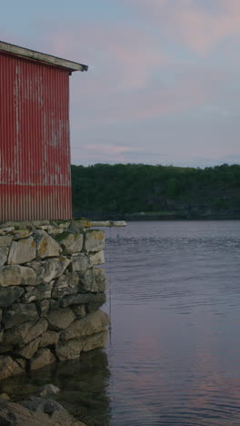a vertical shot of a weathered red boathouse on a rocky shoreline with calm water in a norwegian fjord.