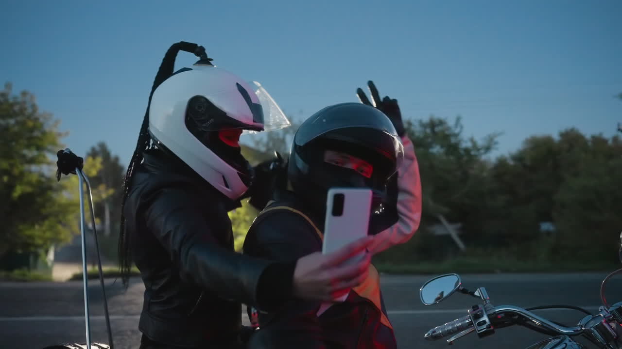 Husband and wife wearing helmets sit on motorcycle during evening, wife extends arm with smartphone taking selfie as husband looks ahead, glowing sunset and street light