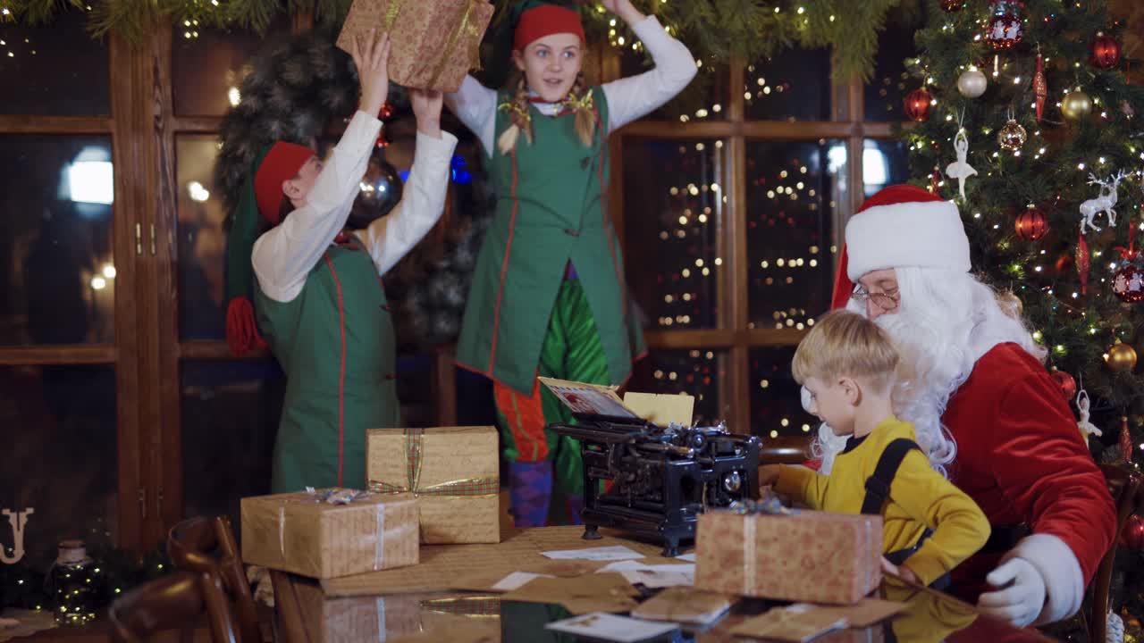 Happy elves playing with boxes near Santa. Santa is typing a letter on old machine near the Christmas tree. Joyful atmosphere at Christmas.
