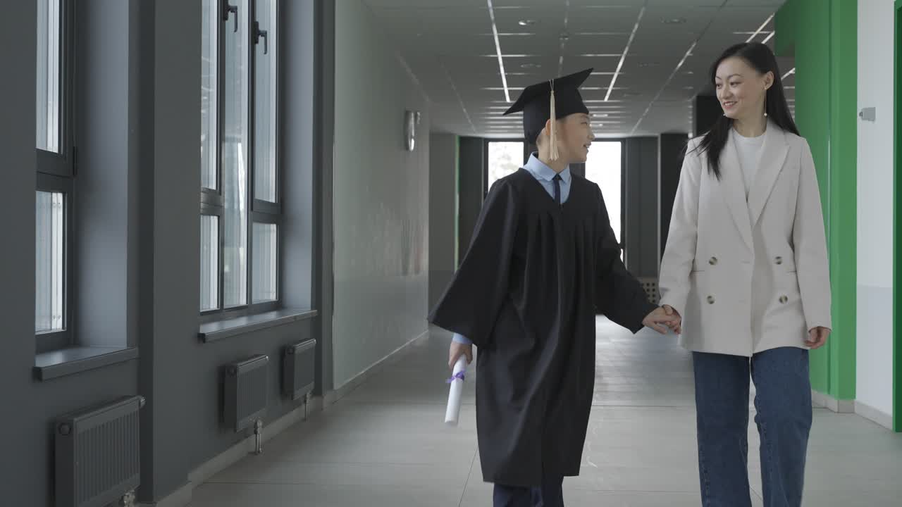 Asian mom hugging and walking with her son. He's wearing a gown and a mortarboard.