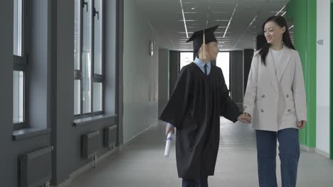 asian mom hugging and walking with her son. he's wearing a gown and a mortarboard.