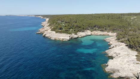 Aerial-view-of-Cala-Escorxada,-the-virgin-beach-in-Menorca,-Spain-with-blue-water-and-a-hill-nearby-with-green-trees-and-blue-clear-sky-in-the-background