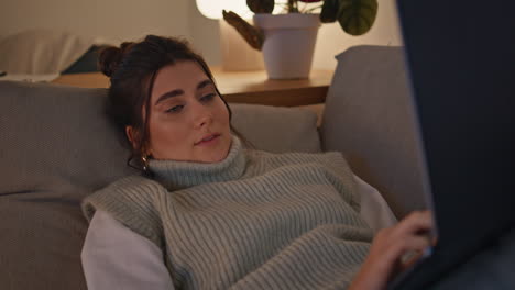 chica tranquila relajándose en el sofá escribiendo en el portátil por la noche en el hogar de primer plano. mujer descansando en el sofa