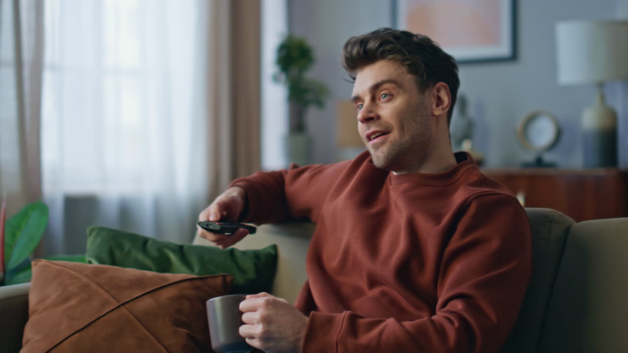 Smilende mand skifter kanaler på tv'et med fjernbetjeningen på sofaen tæt på.