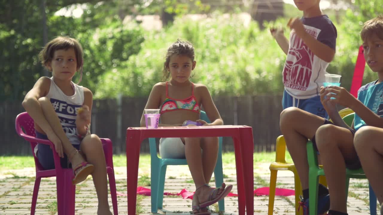 Crianças felizes em um acampamento de verão, sentadas em cadeiras plásticas coloridas, bebendo e comendo lanches, se divertindo ao ar livre em um campo de grama verde, festa infantil em câmera lenta