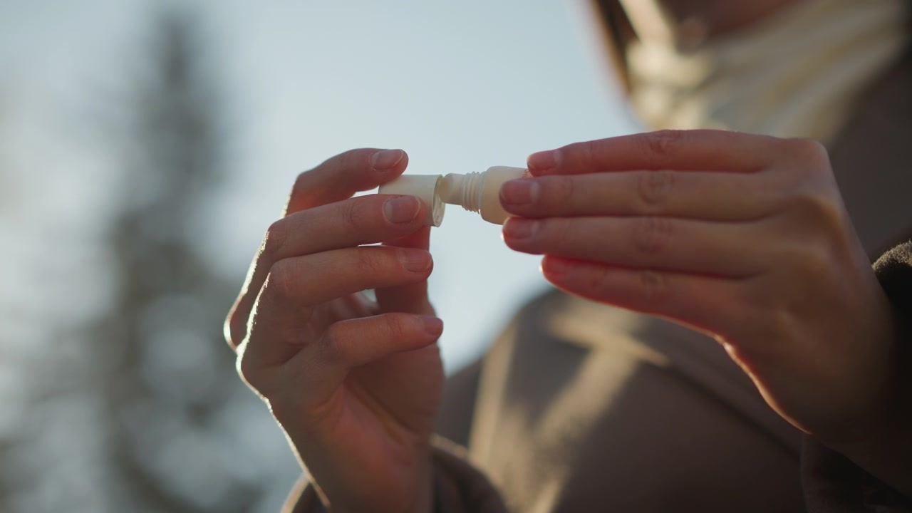 Primo piano delle mani di una persona che aprono e chiudono il tappo di un piccolo tubetto bianco su uno sfondo esterno con illuminazione soffusa. L'attenzione rimane sui movimenti delle mani con il prodotto per la cura personale.