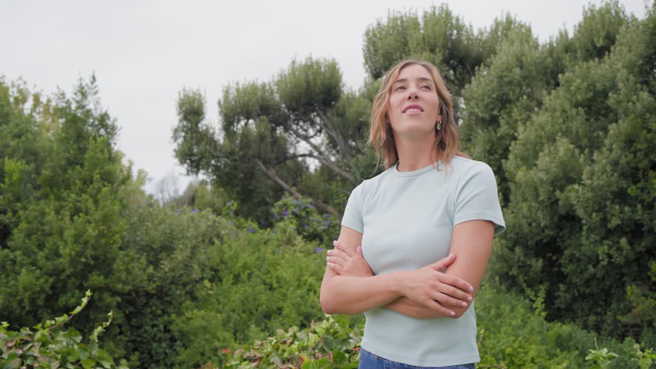 Frau mittleren Alters steht an einer Hecke, blickt auf die Aussicht, dreht sich zur Kamera, sucht Ruhe, in einem petrolfarbenen T-Shirt.