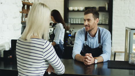 ober in gesprek met blonde vrouw aan de bar, terwijl een serveerster haar koffie schenkt