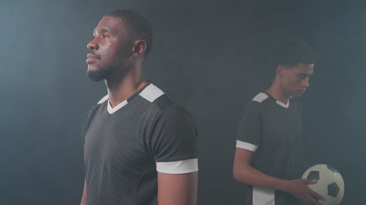 Dúo masculino africano con equipaciones negras en un estudio, el más joven dando un paso adelante con un balón de fútbol, posando.