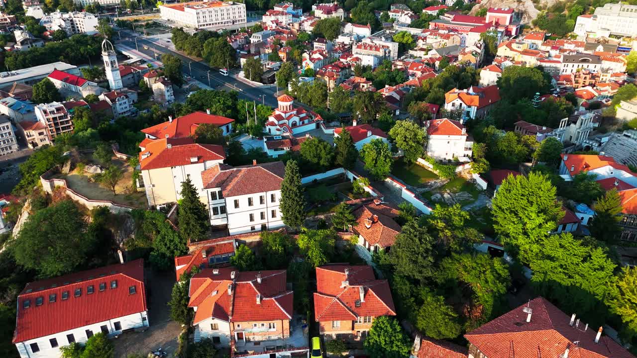 Dron letí nad městem a odhaluje kostel svatého Jiří, amfiteátr, zeleň, střechy a hlavní silnici v teplém světle.