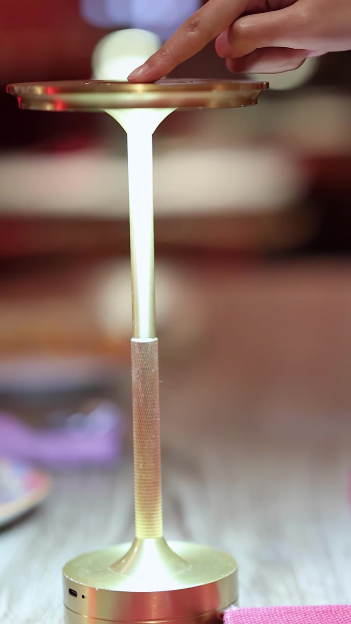 Una mano tocca ripetutamente la parte superiore di una moderna lampada touch dorata, accendendo e spegnendo la sua luce in un ambiente di ristorante illuminato calorosamente