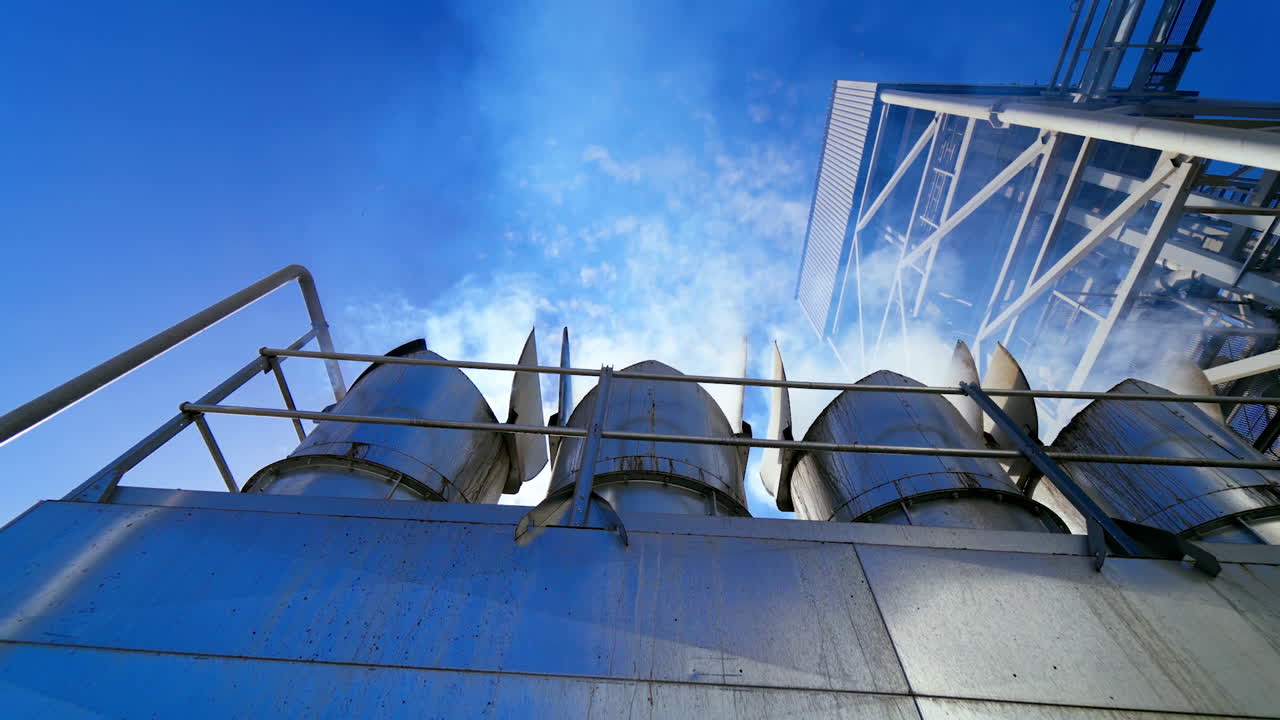 Exterior of warehouse at sunlight. White smoke go into the air from metal pipes of modern granary. Industrial equipment for agribusiness. View from below.