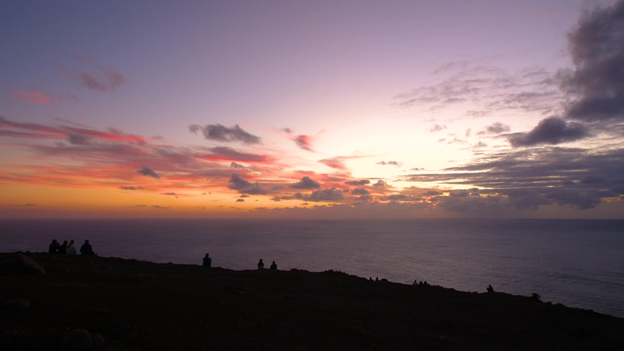 Sunset over ocean horizon, silhouett figures sit calmly in the foreground, enjoying the scenery clouds streak across the darkening sky, with vibrant orange, pink, and sky of purple hues