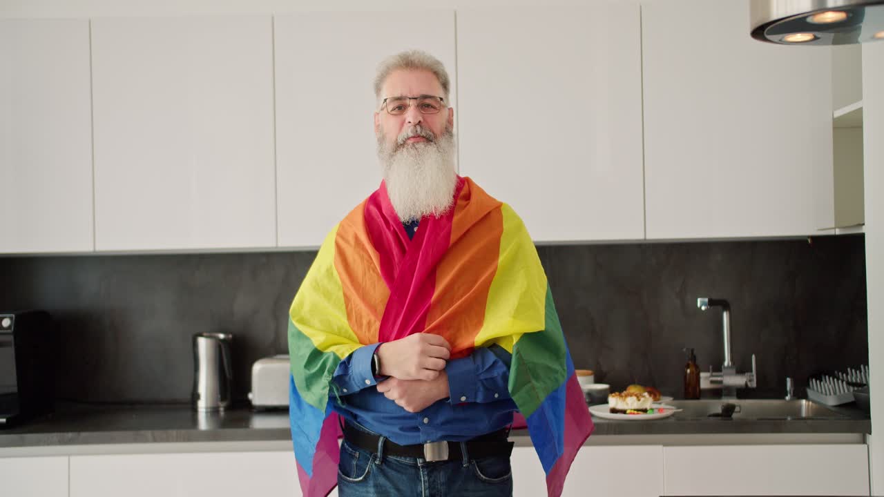 un anciano feliz con cabello gris y una barba exuberante en gafas se levanta por encima de sí mismo y luego envuelve a una persona lgbt en una cocina moderna en un apartamento soleado. retrato de un anciano felice apoya a la comunidad lgbt que posa junto con la bandera lgbt