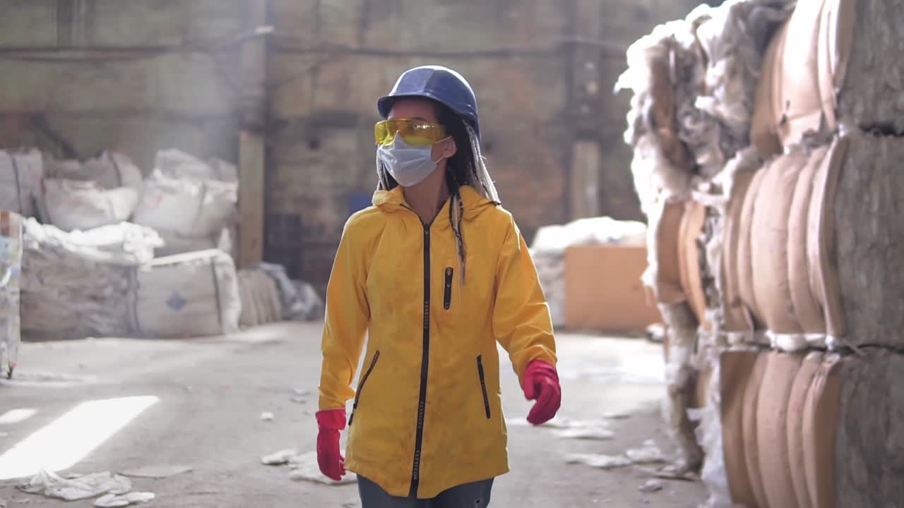Waste processing plant. Recycling and storage of waste for further disposal. Woman worker in hard hat, gloves and mask walking through the stacks of pressed disposal waste, huge piles. Front view footage