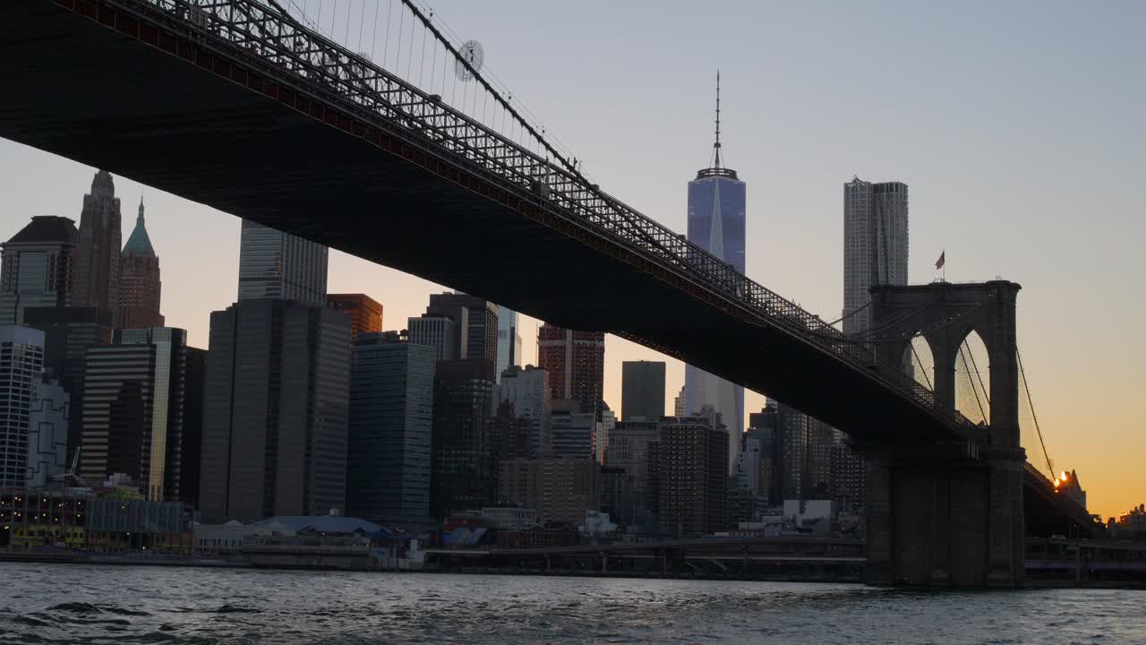 Drone video capturing Brooklyn Bridge silhouetted against Manhattan's glowing skyline at dusk. Water reflections and warm sunset hues create a captivating cityscape