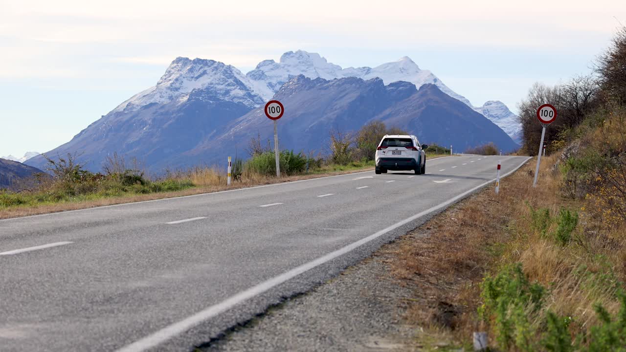 A car travels along a mountain road with stunning views of snow-capped peaks under clear skies
