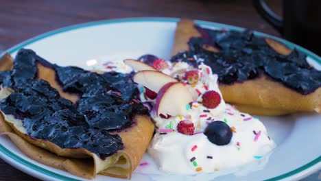 crepas caseras rellenas de queso crema, cubiertas con mermelada de arándanos, servidas con crema batida y frutas frescas como manzanas, fresas y arándanos