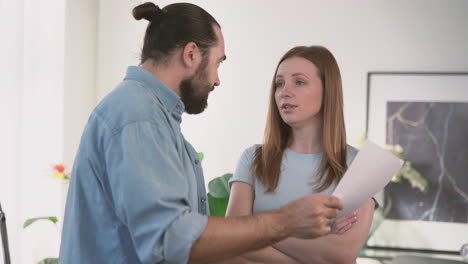 very angry woman asks a bearded man to explain himself to her over some papers