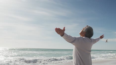 Back-view-of-hispanic-senior-woman-standing-on-beach-and-raising-hands