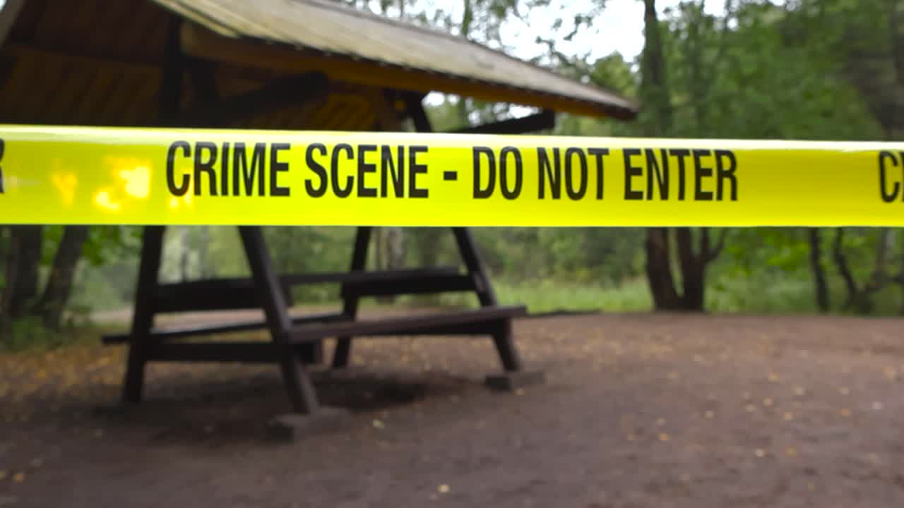 Yellow colored police line Crime Scene forensic investigation tape ribbon in front of a camping site or grilling spot in a forest while the background is bokeh blurry. Fallen leaves on sandy ground
