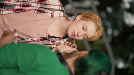 Over-the-shoulder:-Vertical-video-of-a-girl-with-green-hair-in-a-Green-shirt-doing-make-up-to-her-blonde-girlfriend-with-a-short-haircut-in-a-checkered-shirt-during-her-date-in-the-park-in-the-summer