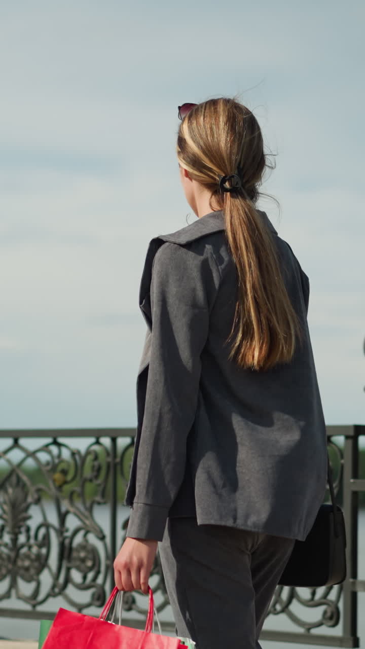 회색 옷을 입은 여자가 장식된 울타리 근처에서 걸어가고, 그녀의 옷이 걸어가는 동안 날아다니며, 그녀의 손에 다채로운 쇼핑 가방의 부분적인 시각으로