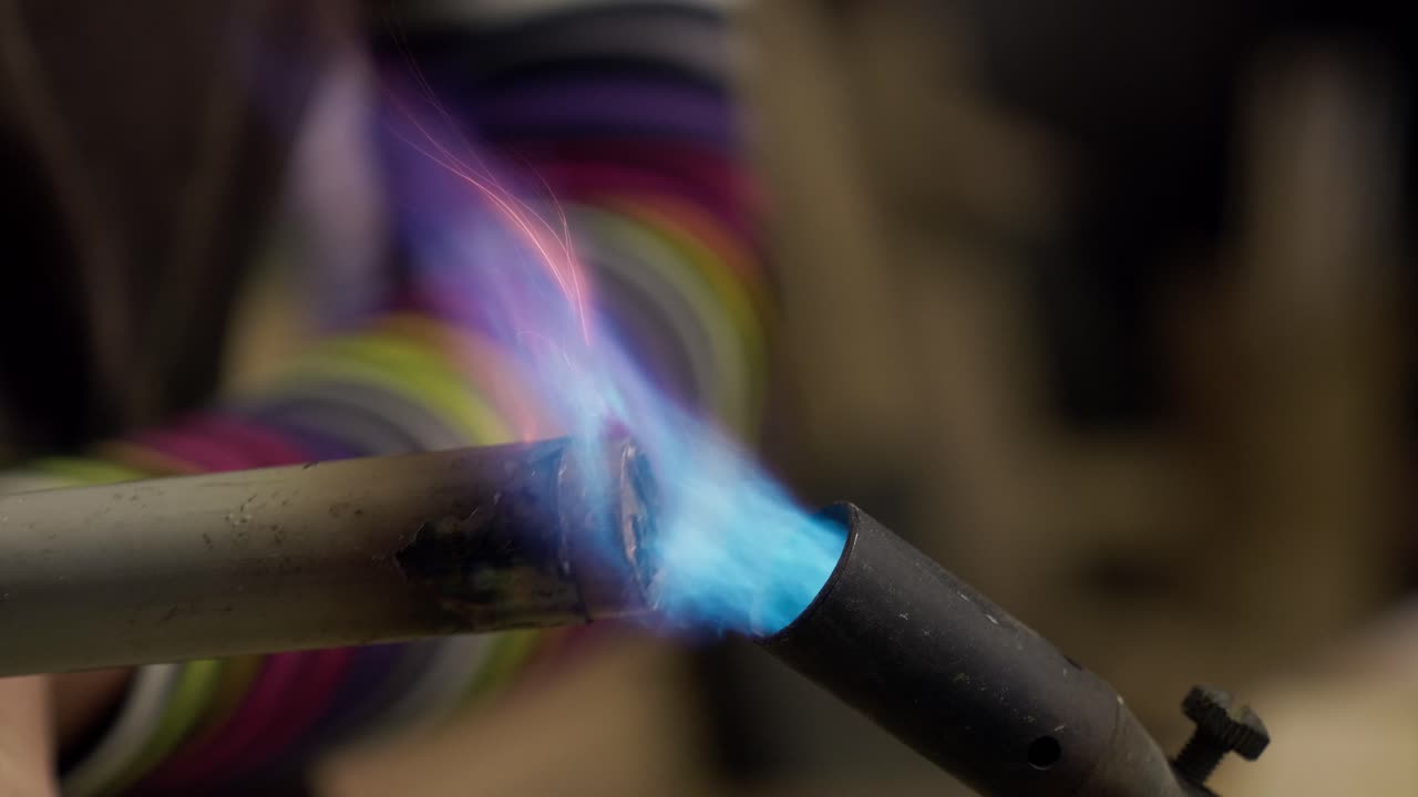 Female craftsperson heating a metal component with a gas torch flame. Industrial process in an artisanal studio. Medium shot, slow motion