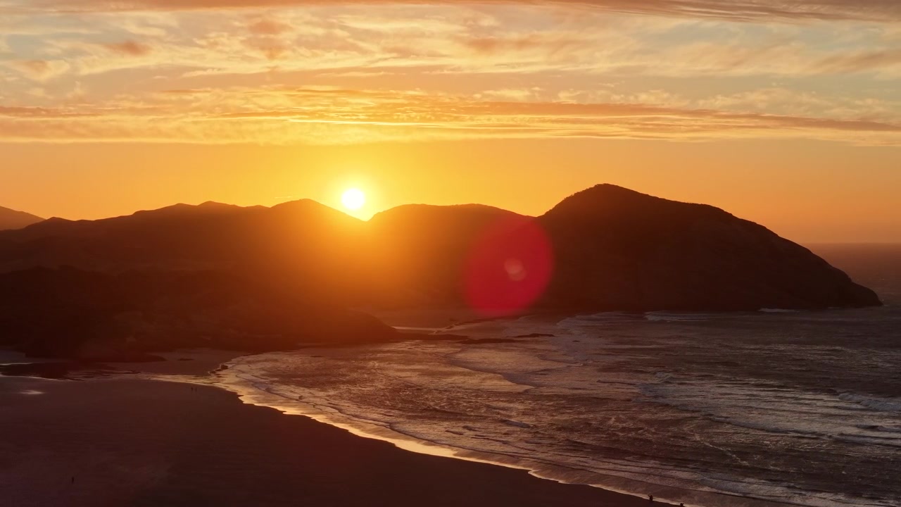 En fantastisk luftudsigt over en strand på New Zealands Sydø ved solnedgang, hvor solen dykker ned bag barske bakker og skaber en levende linseflare over de brændende bølger.