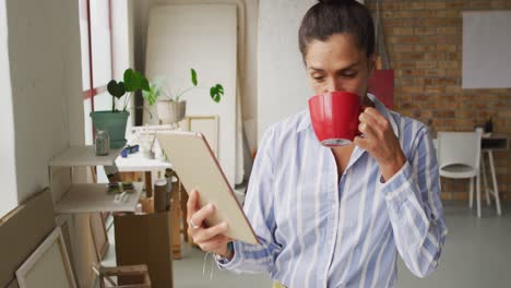 Video-Einer-Glücklichen-Gemischtrassigen-Designerin,-Die-Im-Studio-Ein-Tablet-Benutzt-Und-Kaffee-Trinkt