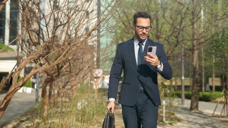 young european business man looking at cellphone having video call while walking on street in modern city park