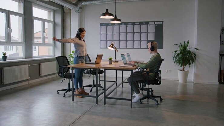 Donna che spinge indietro la sedia, si alza e si stira a una scrivania condivisa, con un laptop visibile.
