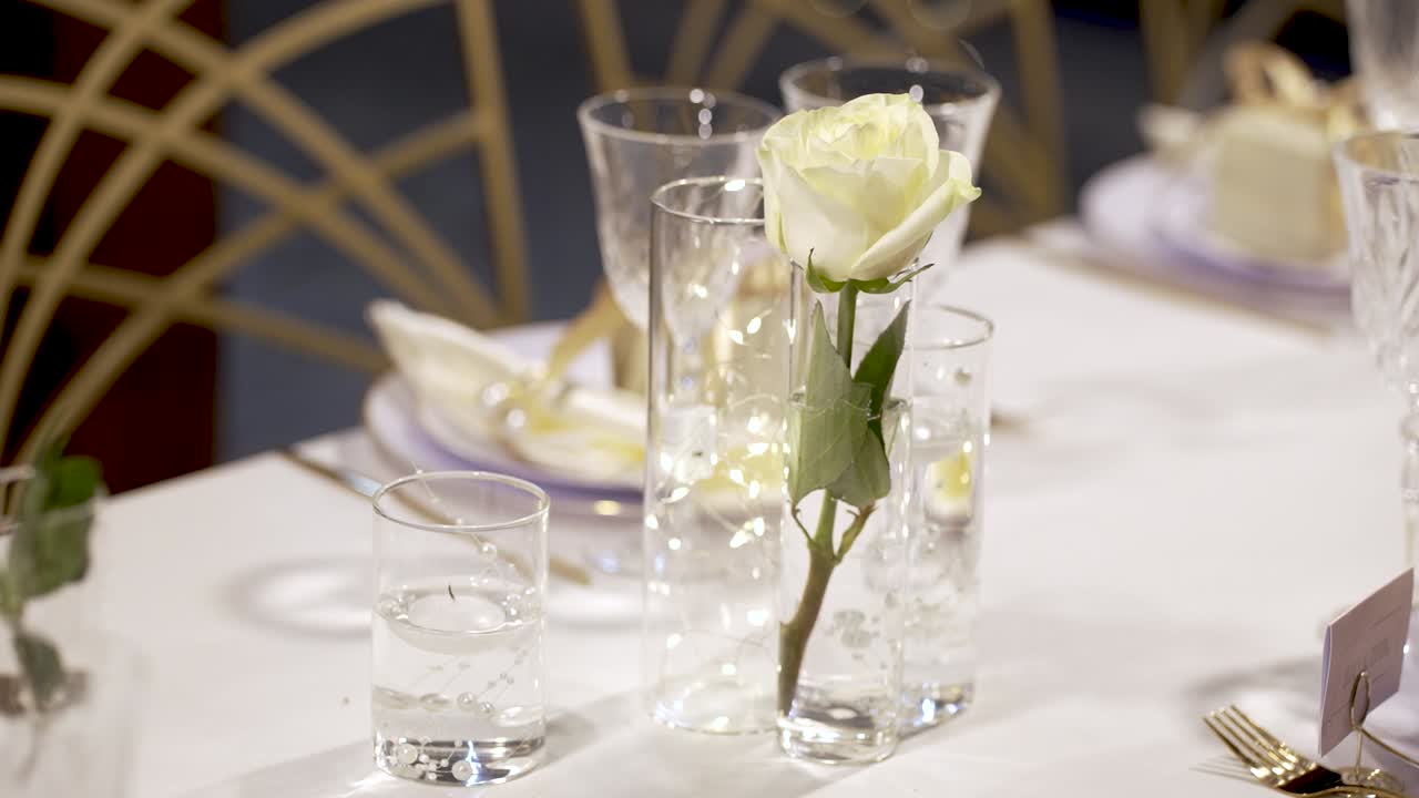 Fresh white roses are placed in glass vases on a beautifully arranged table at a special occasion. The setup showcases sparkling glassware and decorative napkins