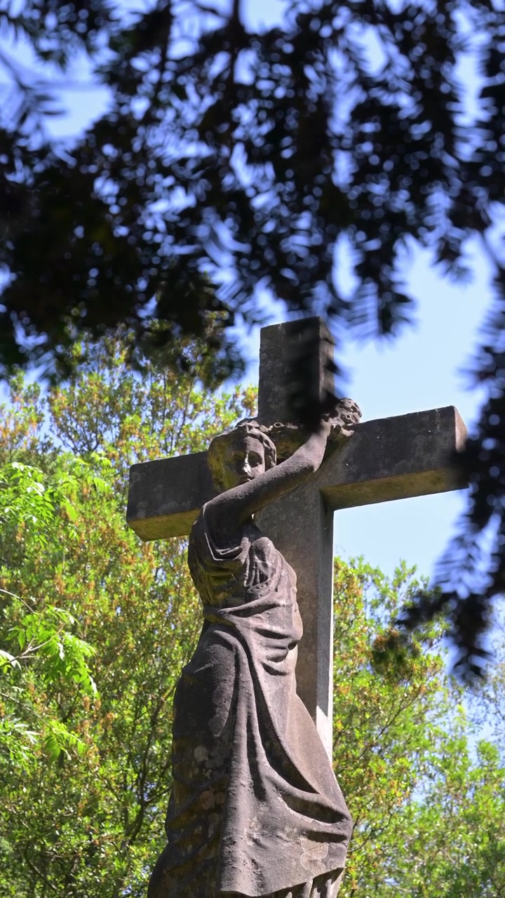 Una statua in pietra consumata dalle intemperie di una figura che tiene una croce si erge tra alberi verdi e lussureggianti. Ideale per documentari storici o contenuti a tema religioso.
