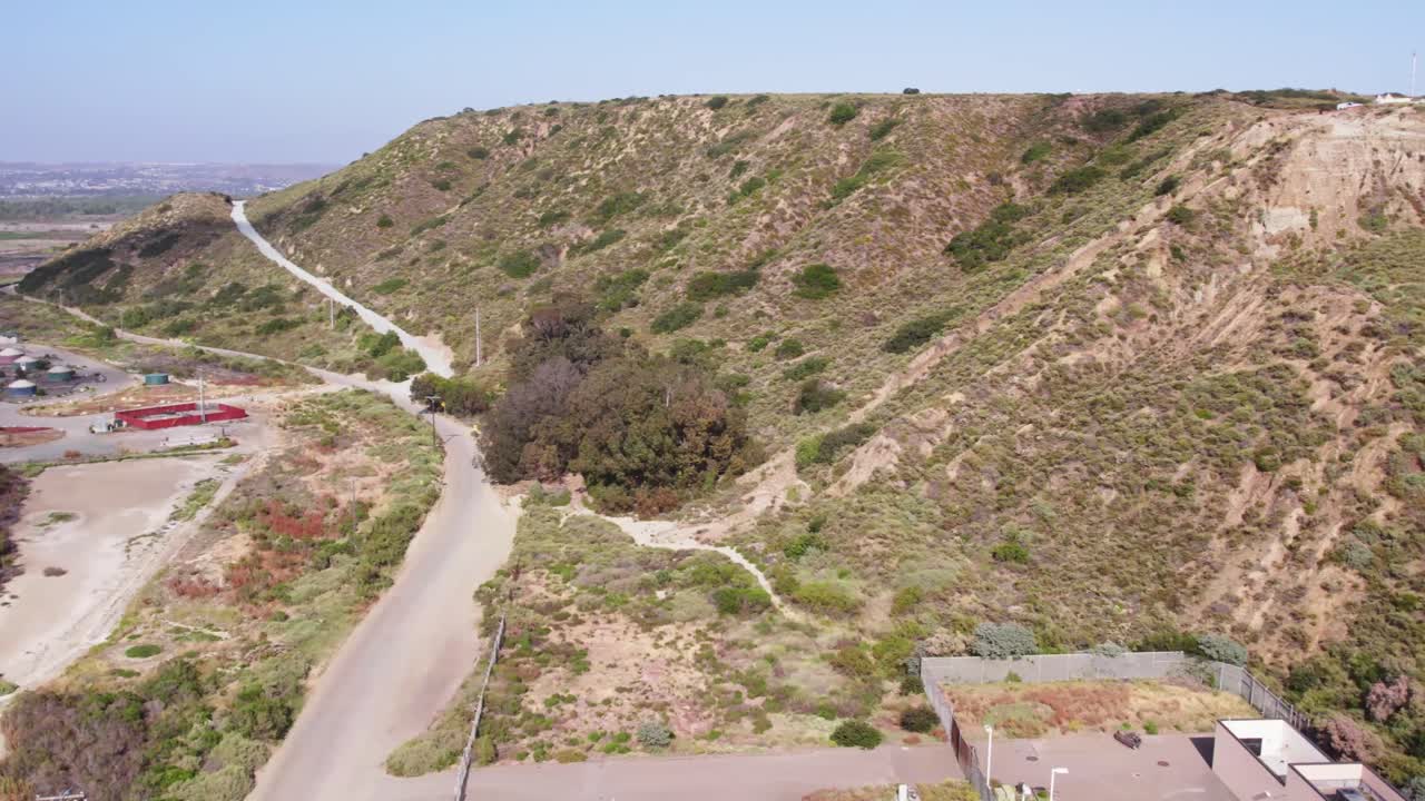 Aerial view of a winding road cutting through rugged desert canyon terrain with dry brush, sparse vegetation, and a security facility near the U.S. borderlands.
