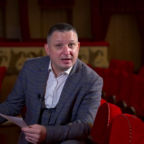 man actor gives interview while sitting on red velvet chair in classic theater. empty theter before evening performance