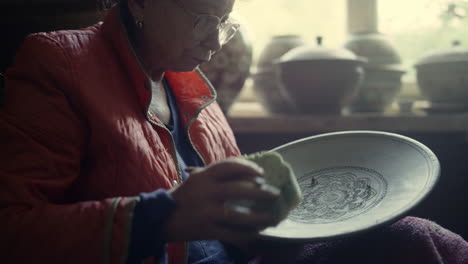 senior ceramist using sponge in studio. woman scraping clay plate in pottery