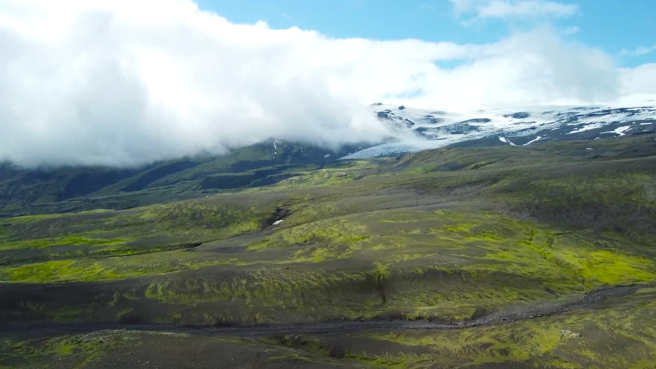 Aerial drone footage flying over green mossy and dark brown rocky Iceland volcanic mountain terrain landscapes during a sunny day with mountains and white vlouds visible in the back, river in front.