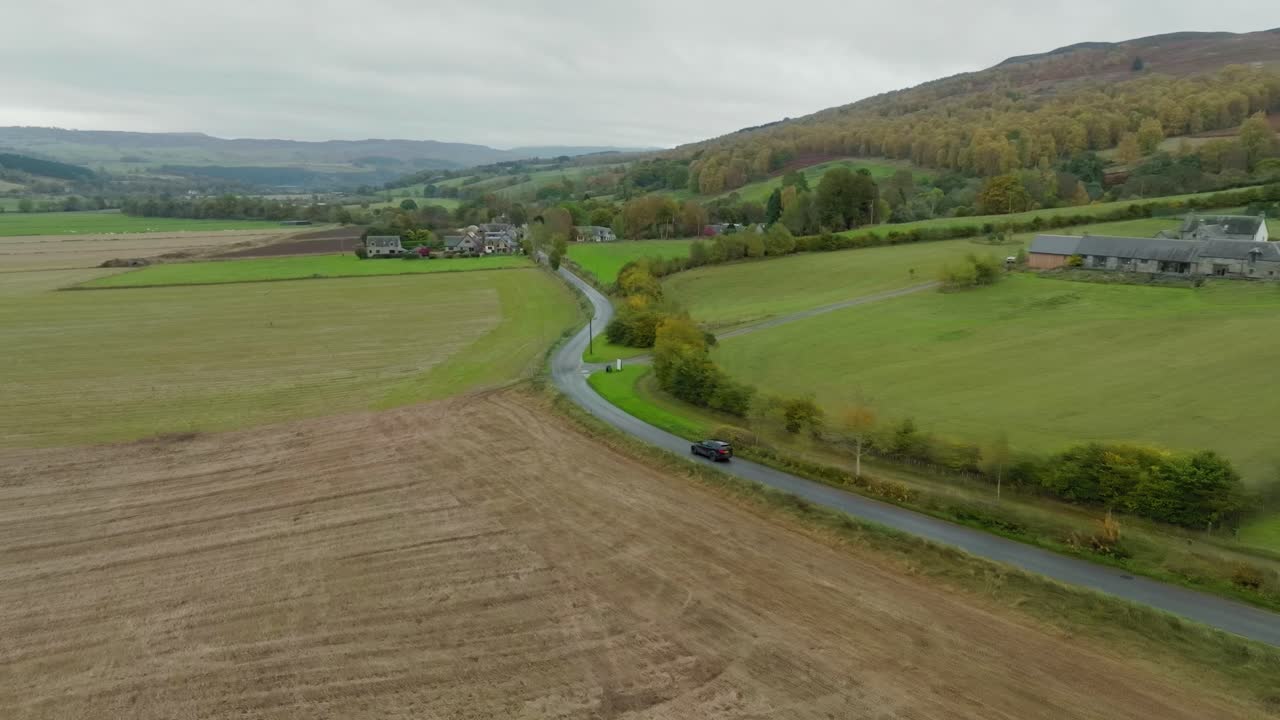 Car driving along countryside road through farmland in Scottish Highland valley and rolling hills, aerial shot over scenic route