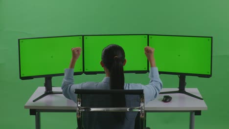 vista trasera de una feliz mujer asiática celebrando mientras trabaja con múltiples monitores de computadora en un estudio de pantalla verde