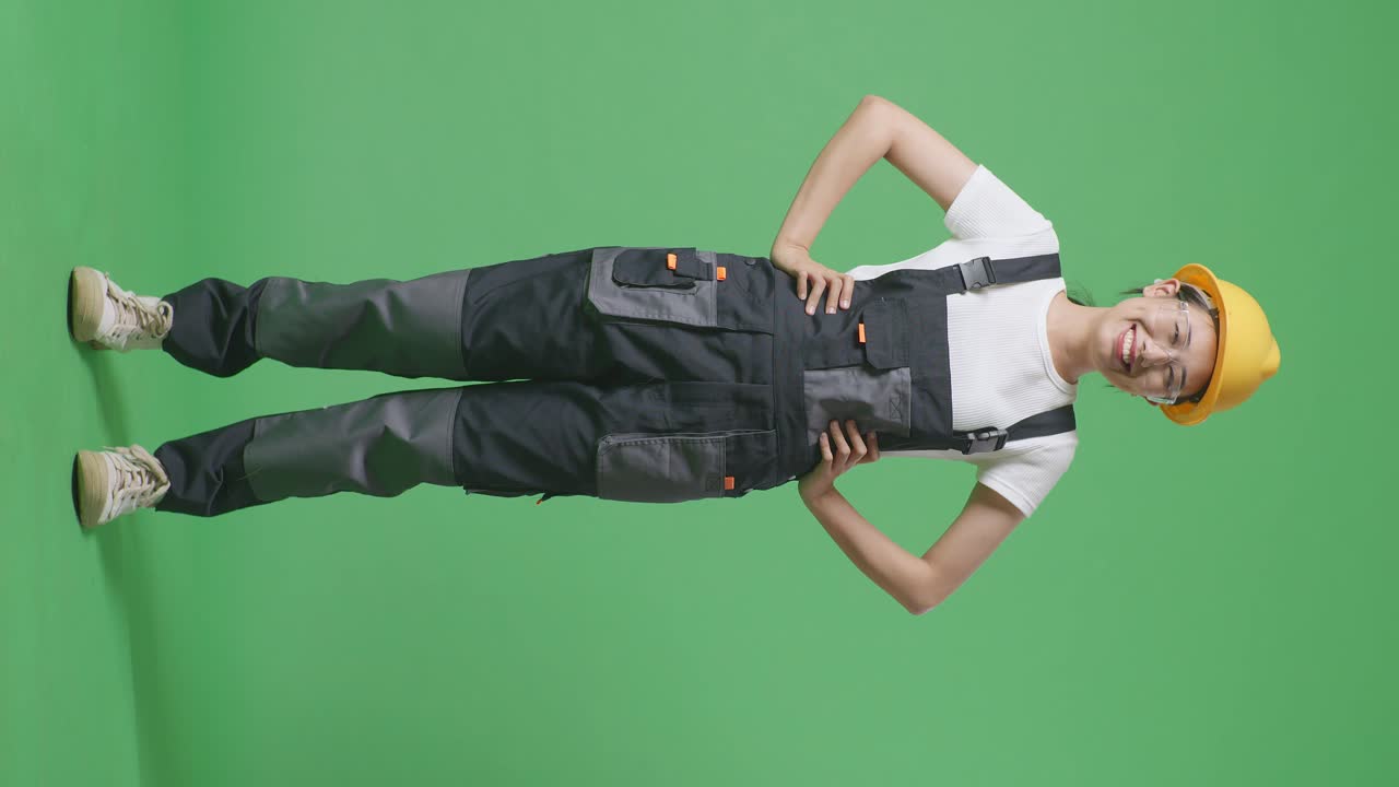 полное тело азиатской женщины рабочая в защитных очках и защитном шлеме стоит с руками акимбо смотрит в камеру и улыбается в зеленом экране фоновой студии