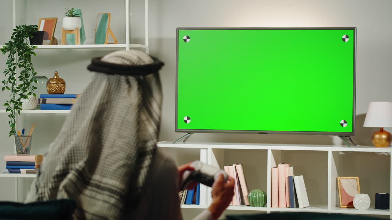 Middle eastern man playing video game, tv with chroma key. Television with green screen, play gaming on modern console. Wearing traditional Islamic male clothes