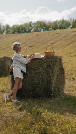 la señora toma fotos de su elegante amiga de pie al lado de la bala de heno con la configuración de picnic en el campo pintoresco, la luz del sol destaca la atmósfera rural relajada