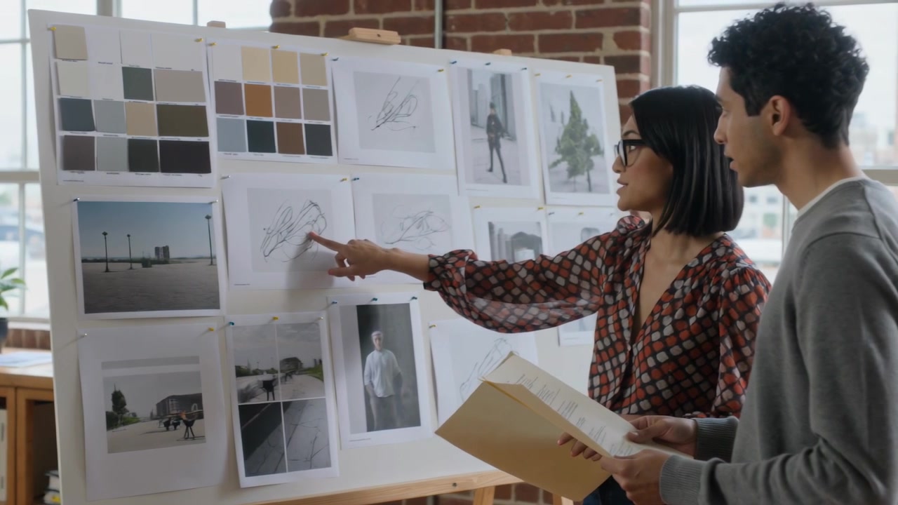 Pointing woman in blouse reviewing board sketches with man holding tan folder in studio, copy space