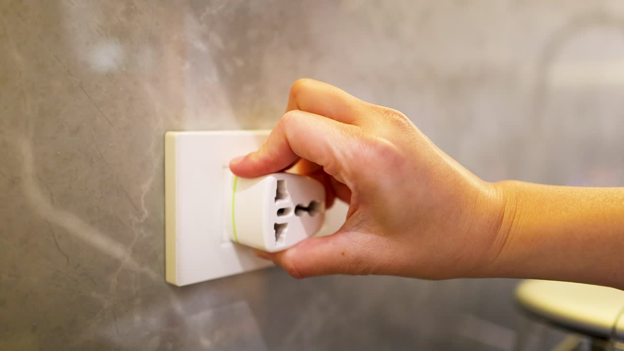 A close-up shot shows a hand removing a white universal travel adapter from a wall