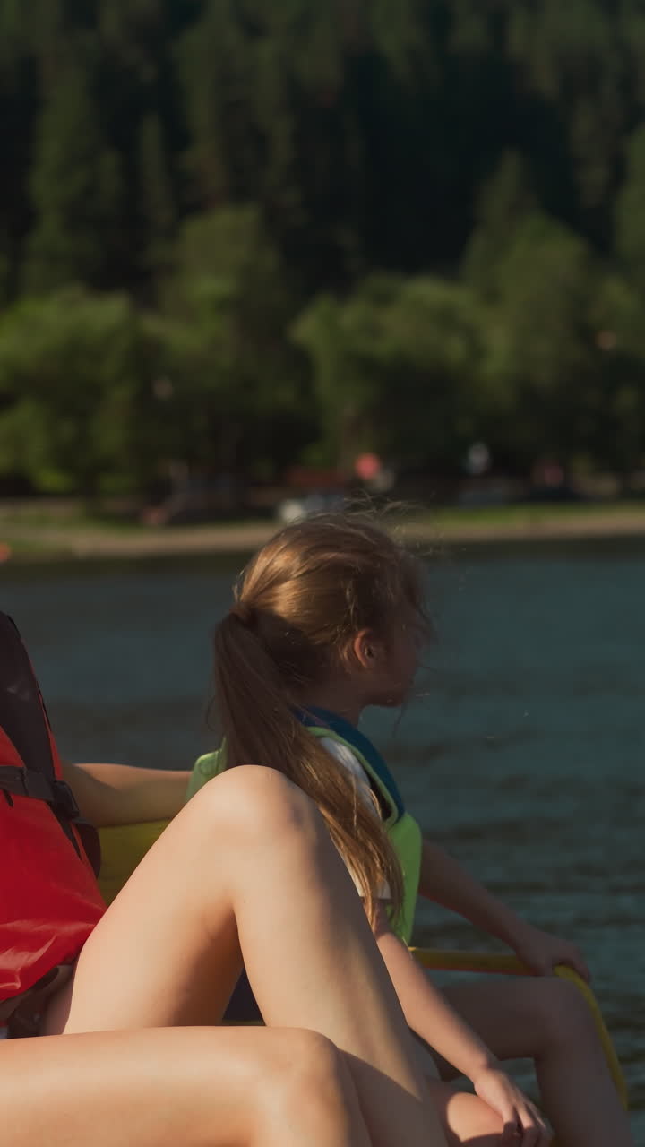 Mother and daughter look at shore with tall trees moving in catamaran. Young woman and little girl in protective vests chat admiring scenery from water transport slow motion