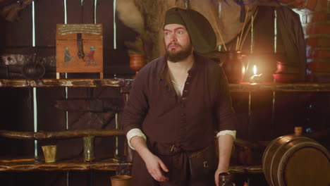 a medieval man in a brown tunic and a green hat stands in a rustic, candle lit room.