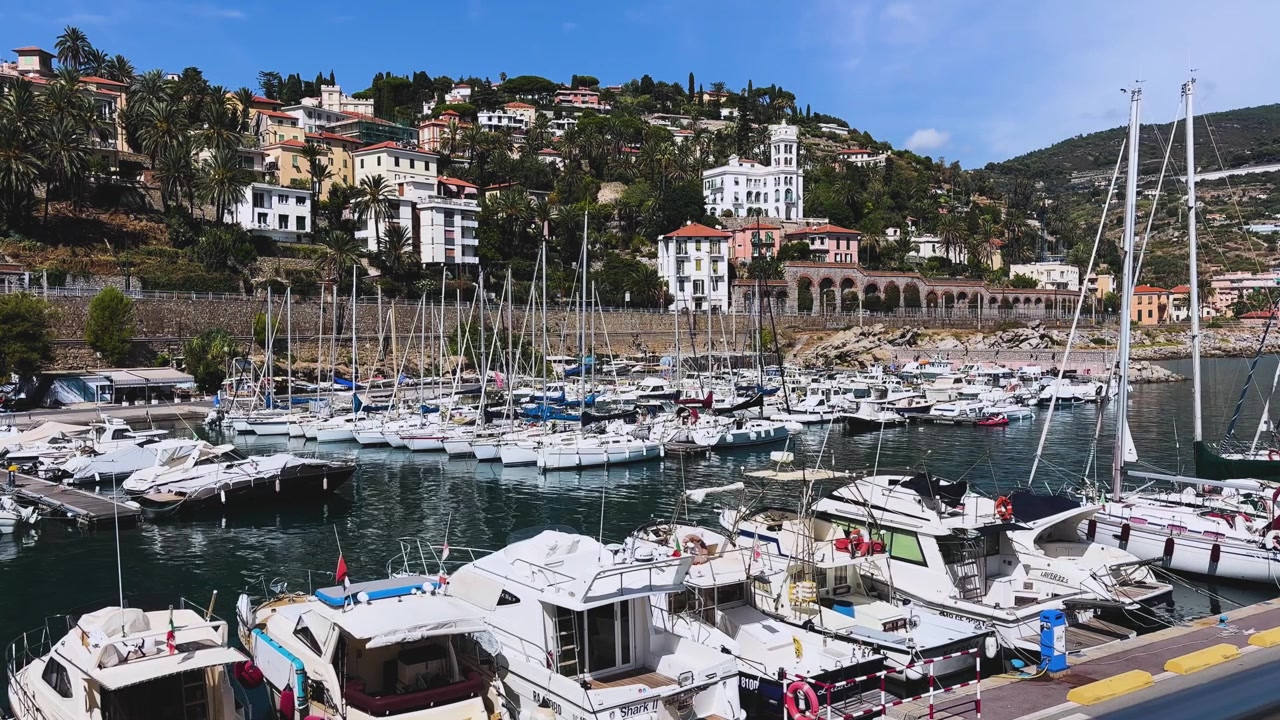 Vista panoramica del Porto di Bordighera, pieno di yacht e barche a vela, con sullo sfondo portici storici, palme e ville collinari sotto un cielo azzurro brillante.