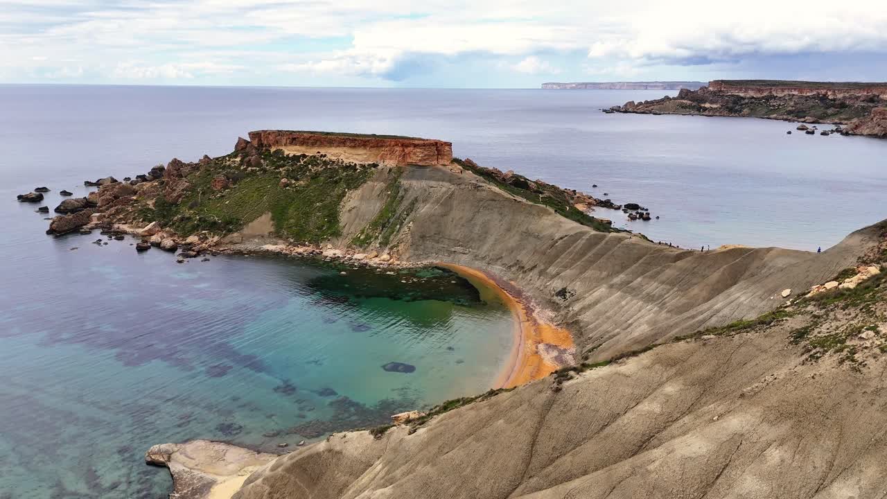 Vista de drone da Baía de Qarraba em Malta com falésias rochosas íngremes, uma pequena praia curva e mar turquesa claro ao longo da costa acidentada.