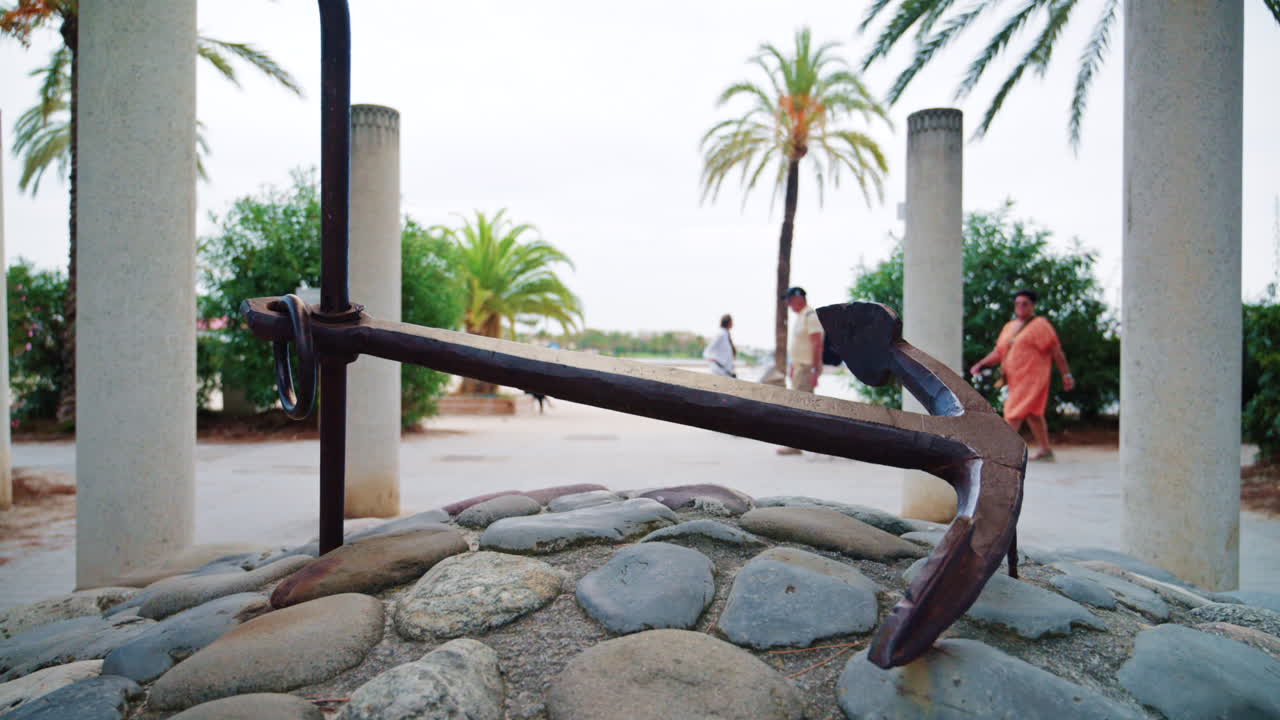 A metal anchor displayed on a stone base near palm trees and a seaside promenade